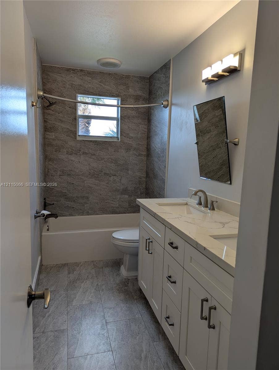 215 SW 120th Avenue, #215, Pembroke Pines, Pembroke Pines, FL 33025 Bathroom featuring a dual-sink vanity with a stone-patterned countertop, grey stone-finish wall tile in the shower area, matching grey tile flooring, and a multi-bulb vanity light fixture