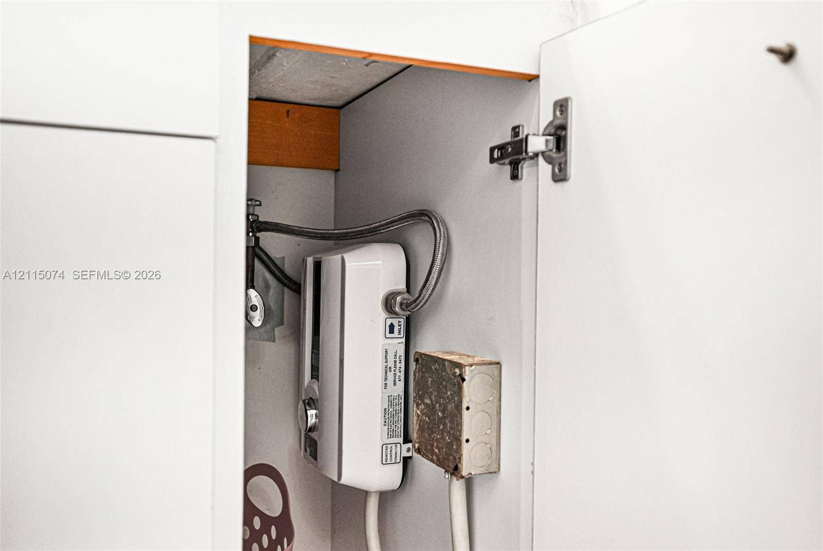 Tankless electric water heater housed within white cabinetry featuring metal braided supply lines and an external electrical junction box