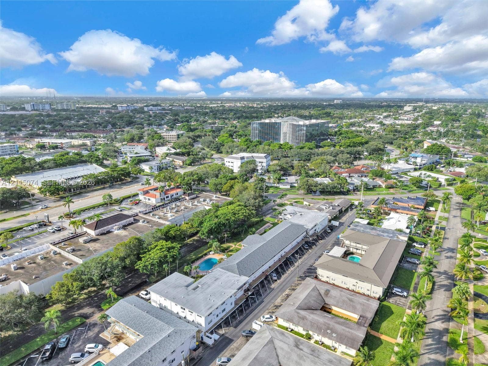 Aerial perspective of the residential complex featuring multi-story buildings, shingled roofing, on-site swimming pools, and manicured landscaping with palm trees