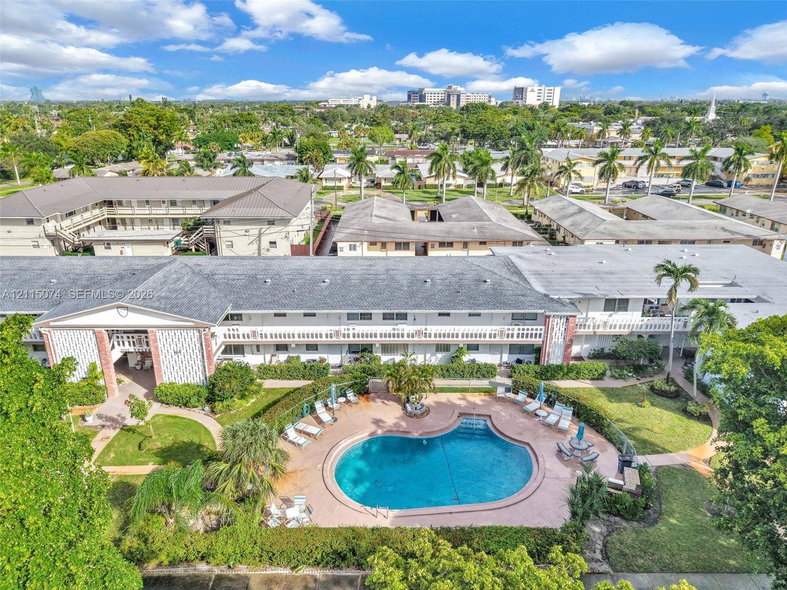 Aerial perspective of a multi-story residential complex featuring a central swimming pool, paved pool deck, manicured landscaping with palm trees, and structural brick accents