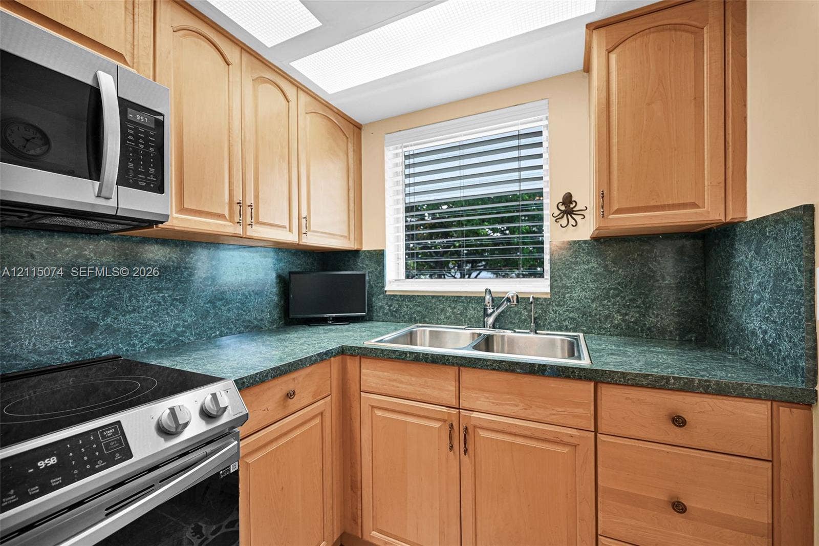Kitchen featuring light-finish wood cabinetry, integrated green stone countertops and backsplash, stainless steel double-basin sink, and built-in stainless steel microwave and oven