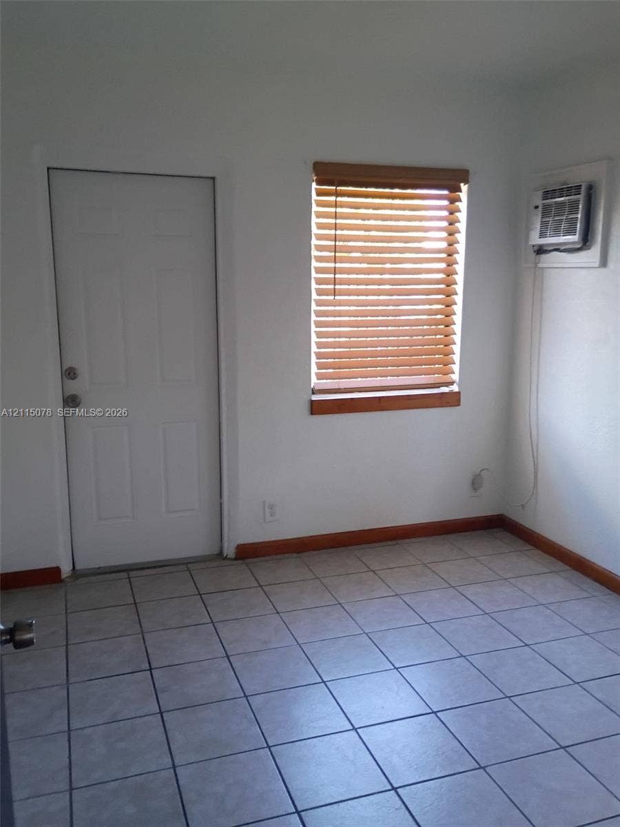 209 SW 12th Street, #A, Dania, Dania Beach, FL 33004 Light-filled room featuring neutral tile flooring, a window with wood-finish blinds, an exterior-access door, and a wall-mounted climate control unit