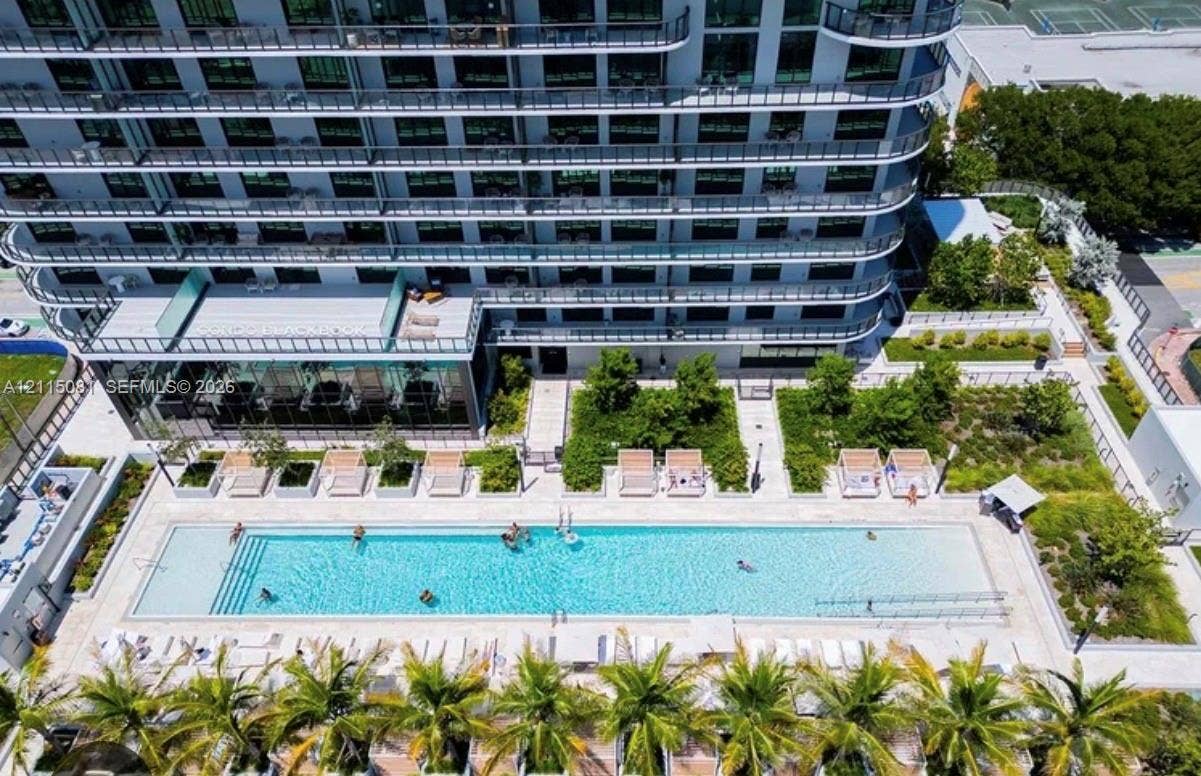 580 72nd Street, #1104, Miami Beach, FL 33141 High-rise residential exterior featuring a multi-story glass facade with private balconies, a rectangular swimming pool, integrated landscaping with mature palm trees, and paved outdoor lounging areas