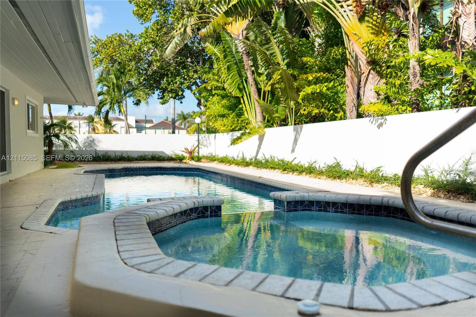 663 Woodcrest Road, Key Biscayne, Key Biscayne, FL 33149 In-ground swimming pool and attached spa featuring decorative tile accents, light-toned stone coping, surrounding hardscape patio, and a privacy wall