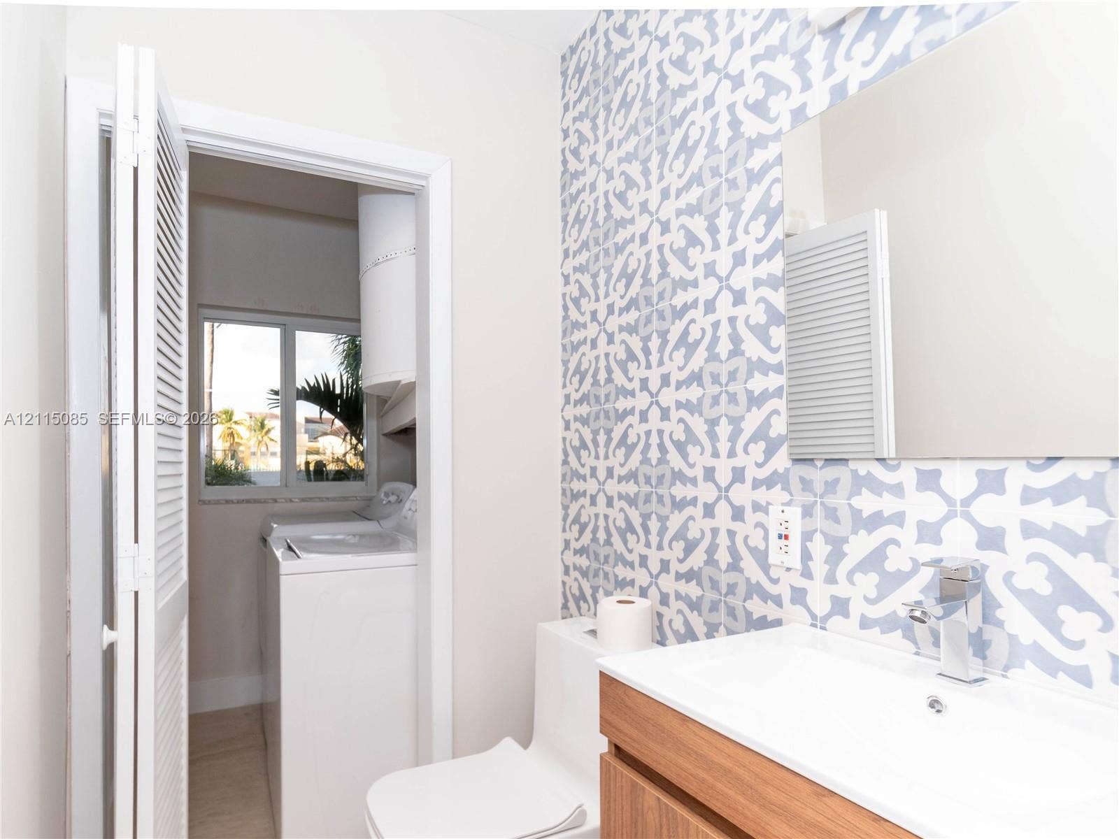 663 Woodcrest Road, Key Biscayne, Key Biscayne, FL 33149 Bathroom featuring a wood-finish vanity with a rectangular sink, wall-mounted frameless mirror, patterned tile accent wall, and a louvered closet door concealing laundry hookups