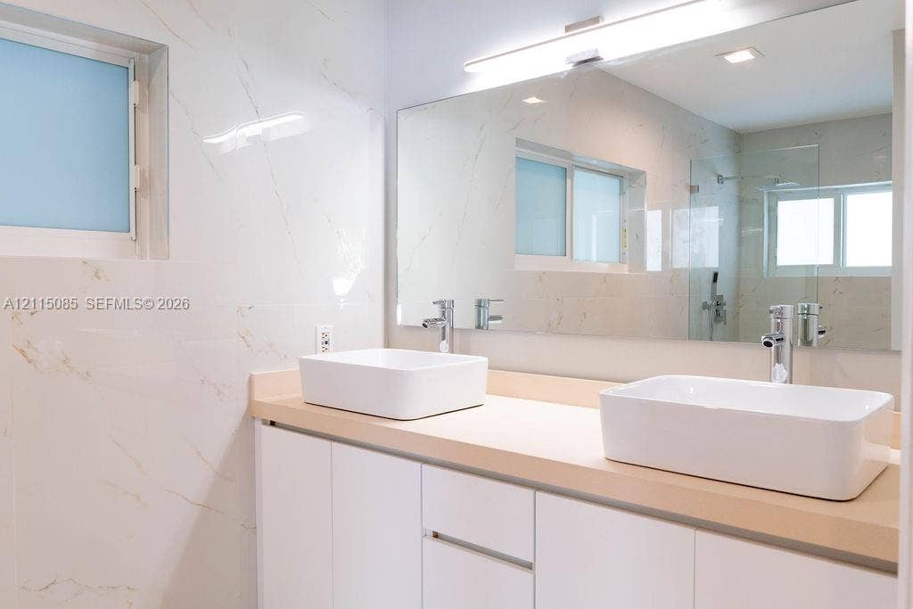 663 Woodcrest Road, Key Biscayne, Key Biscayne, FL 33149 Double vanity featuring white vessel sinks, polished chrome faucets, a solid-surface countertop, white cabinetry, and wall-to-wall mirror with integrated linear lighting, all accented by large-format, veined tile wall finishes