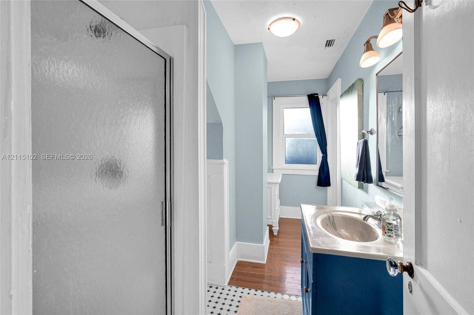 110 S Peninsula, New Smyrna Beach, FL 32169 Bathroom featuring a vanity with an integrated basin, a walk-in shower with a framed glass door, wood-finish flooring, a mosaic tile floor section, and wall-mounted light fixtures