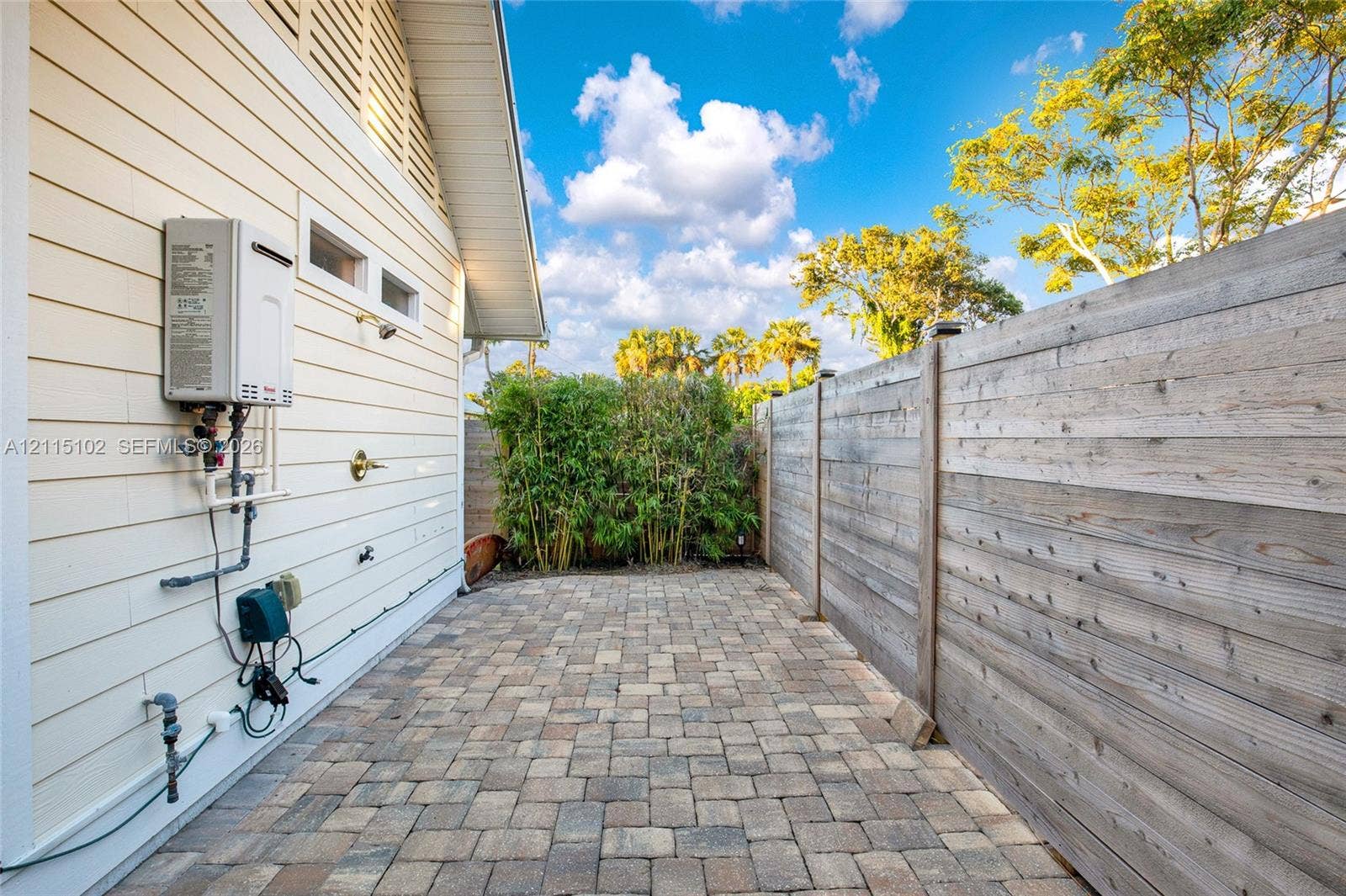 110 S Peninsula, New Smyrna Beach, FL 32169 Exterior side patio featuring light-colored horizontal lap siding, brick paver flooring, a wall-mounted tankless water heater, an outdoor shower fixture, and a wood privacy fence
