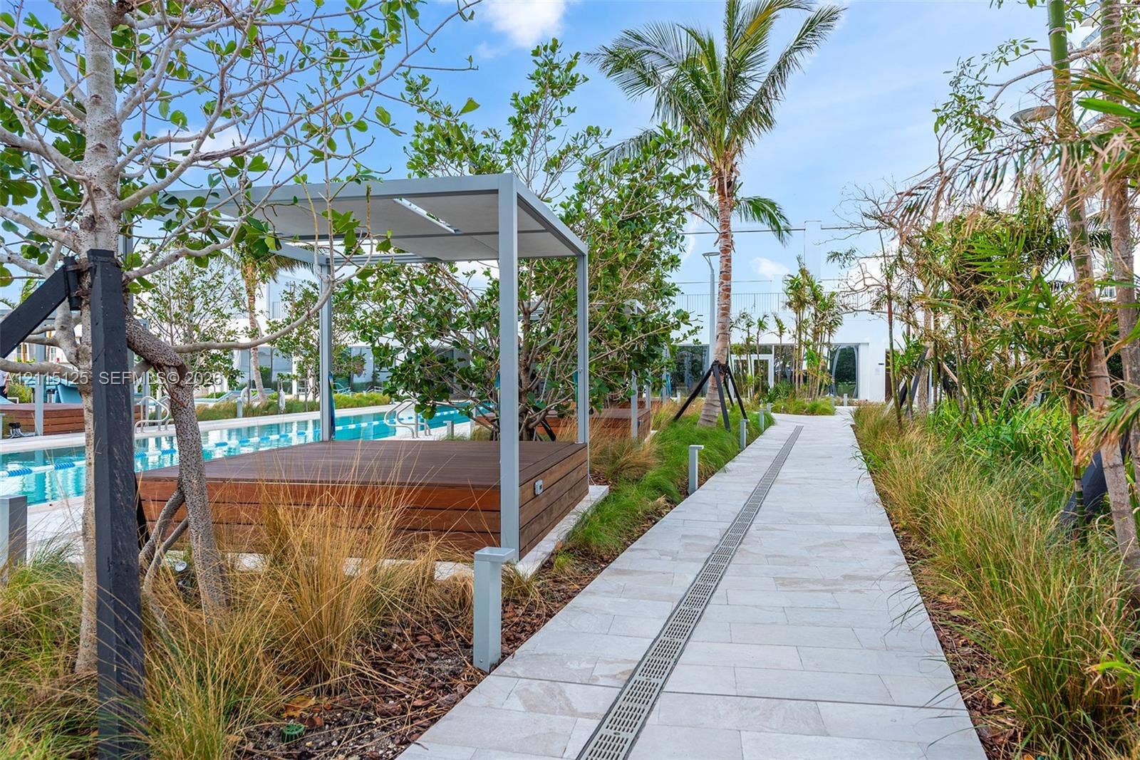 Outdoor lounge area featuring a wood-deck platform with a modern metal-frame canopy, stone paver pathway, and mature tropical landscaping