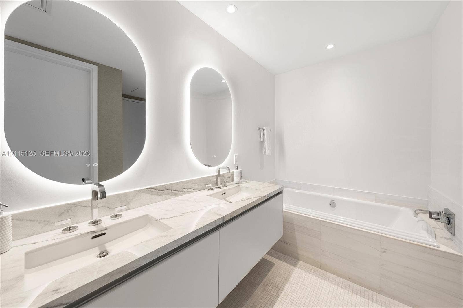 Floating dual-sink vanity with a solid surface countertop, backlit oval mirrors, and a deep soaking tub featuring tiled surround