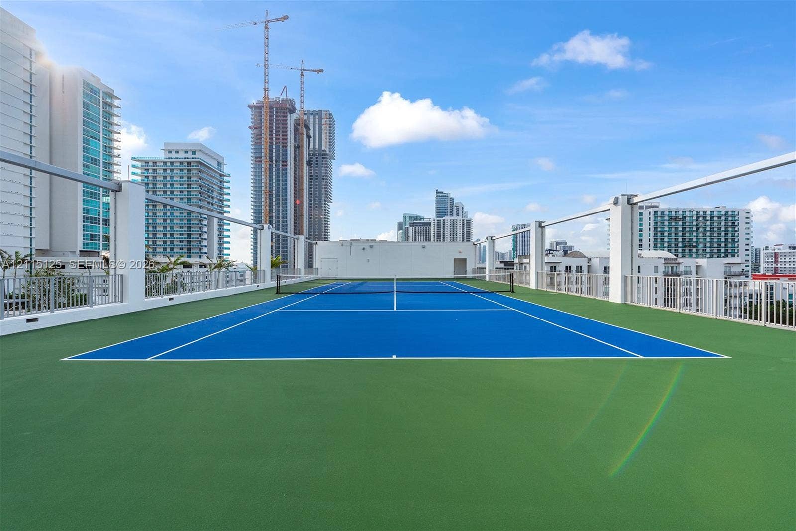 Rooftop tennis court featuring a blue surface with white court markings, green perimeter surfacing, and a white metal perimeter railing