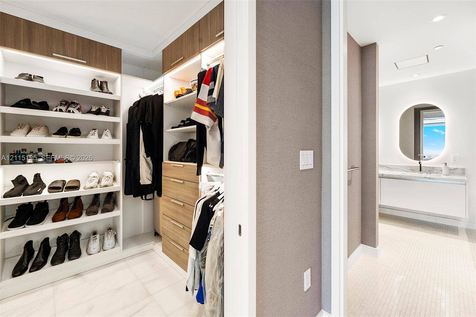 Walk-in closet featuring custom wood-finish cabinetry, built-in shelving with integrated lighting, and a multi-drawer dresser unit, paired with a bathroom showcasing a floating vanity, stone-surface countertop, and backlit oval mirror