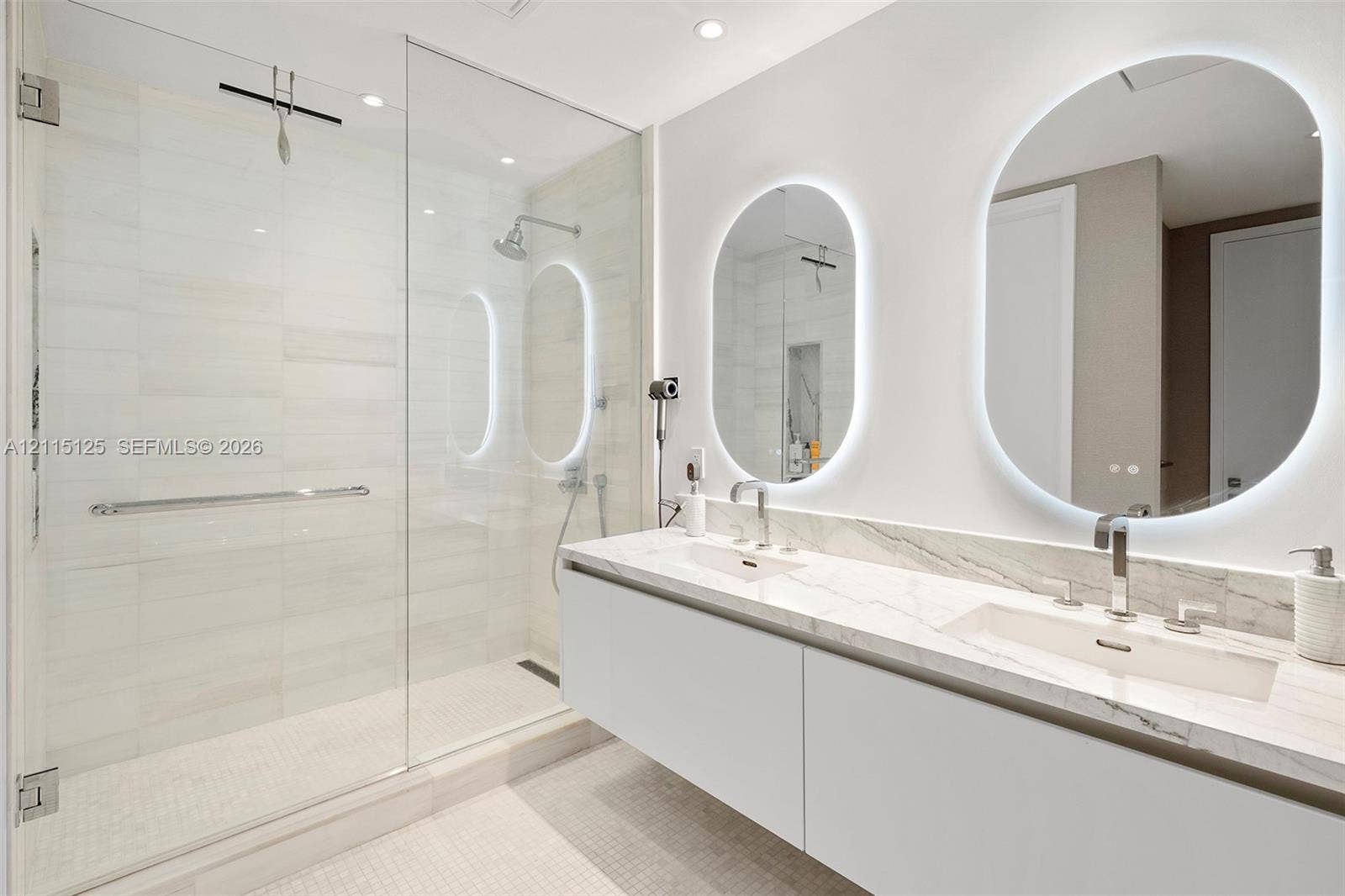 Floating dual vanity with stone countertop, integrated sinks, chrome fixtures, backlit oval mirrors, and a glass-enclosed shower featuring tile surrounds