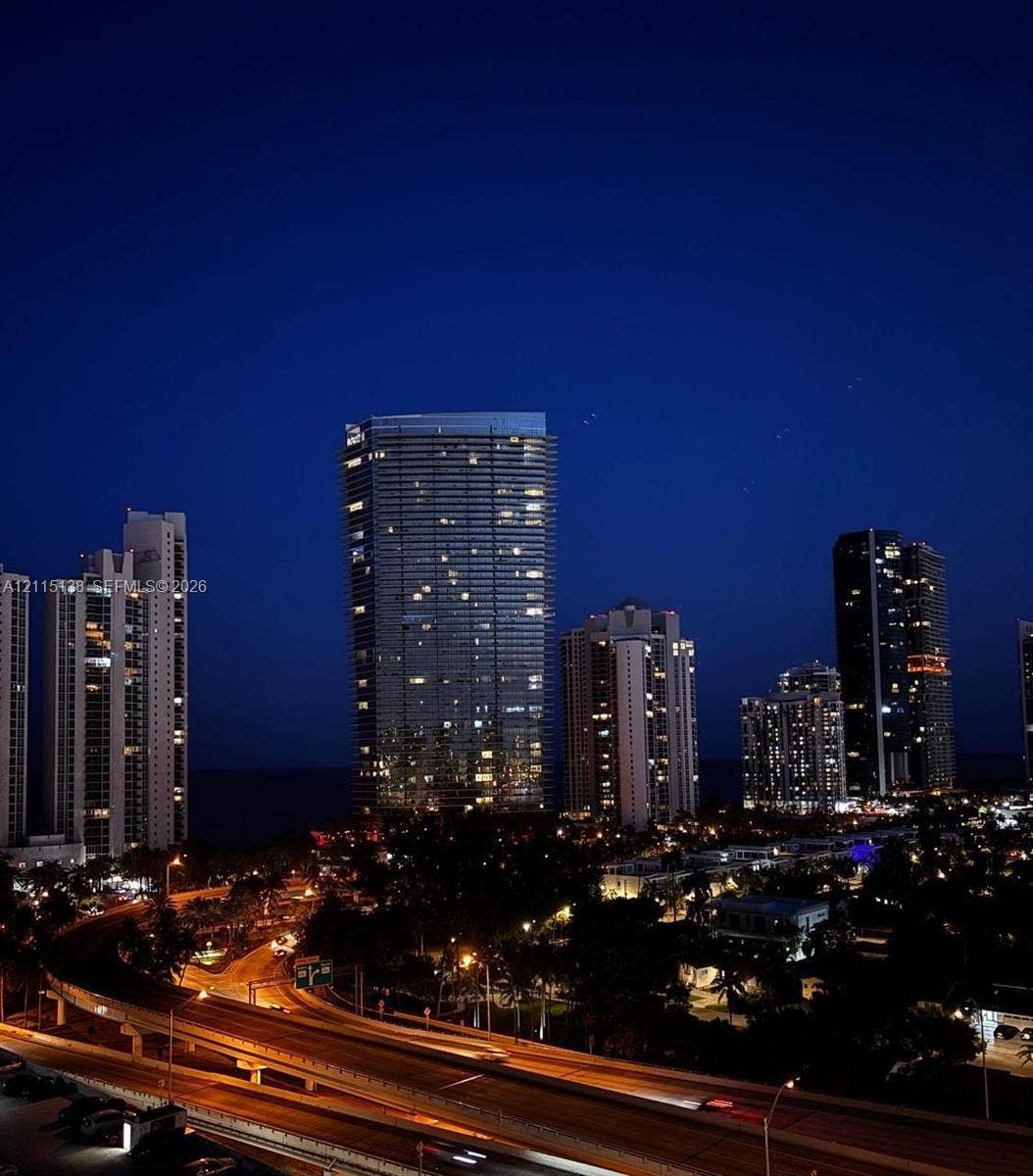 19380 Collins Avenue, #1602, Sunny Isles Beach, FL 33160 High-rise urban landscape featuring illuminated skyscrapers, expansive coastal water horizon, and infrastructure including multi-lane roadways with street lighting