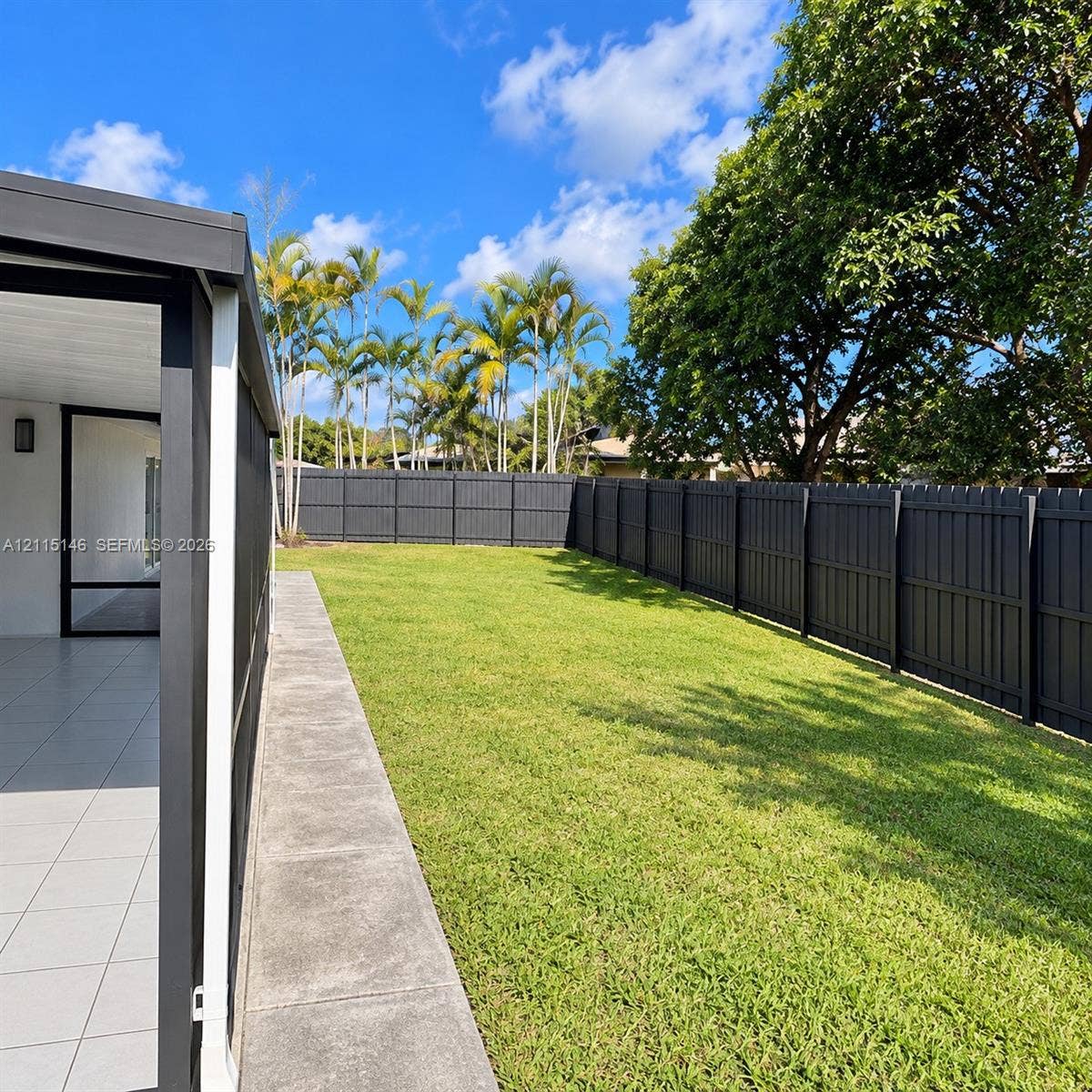 Enclosed patio with tile flooring and a grass backyard featuring a dark wood-finish privacy fence and mature palm trees