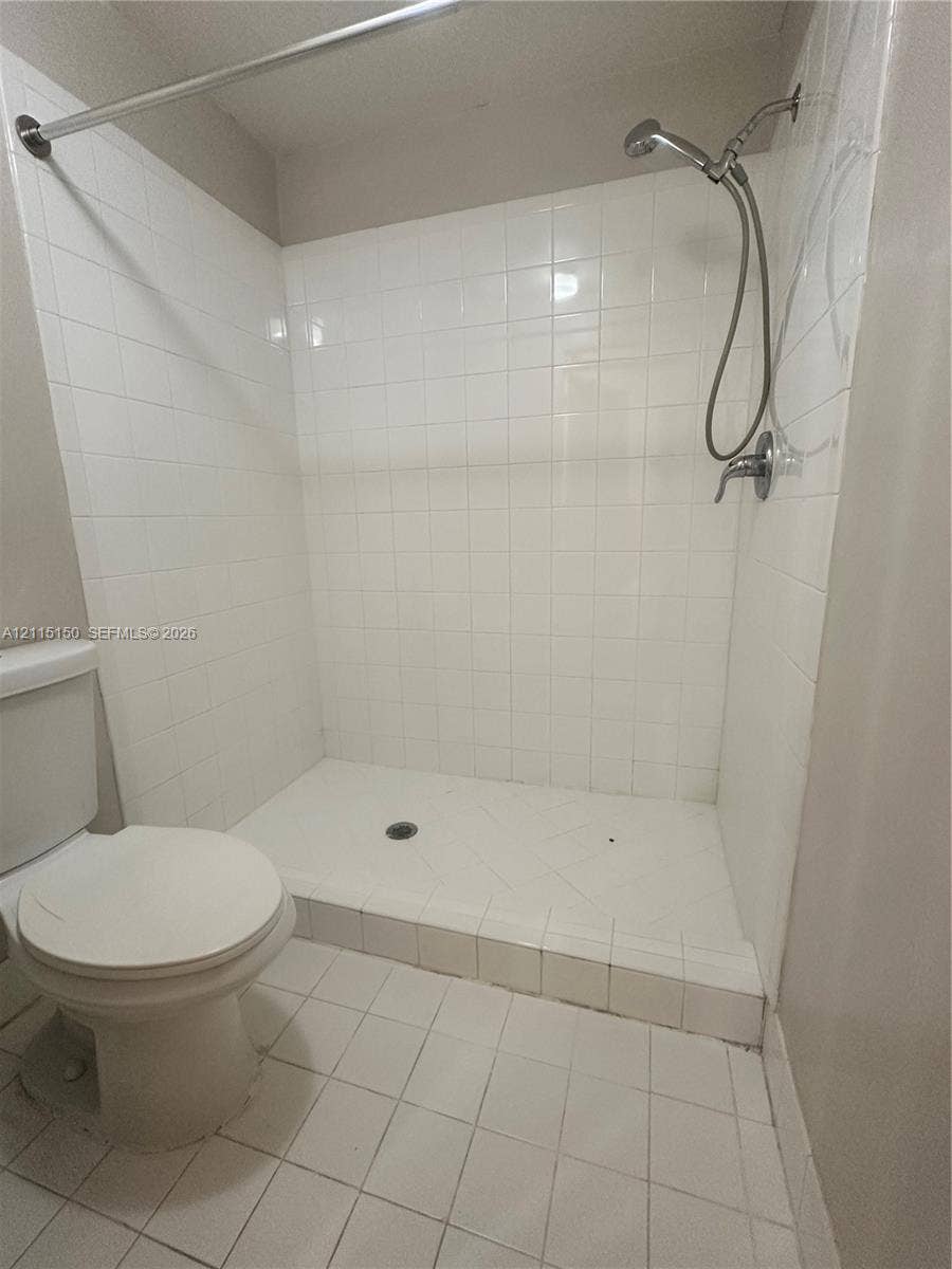 6992 SW 39th Street, #I201, Davie, Davie, FL 33314 Walk-in shower enclosure featuring white wall tiling, a raised tiled threshold, and a chrome handheld shower fixture, accompanied by a white porcelain toilet