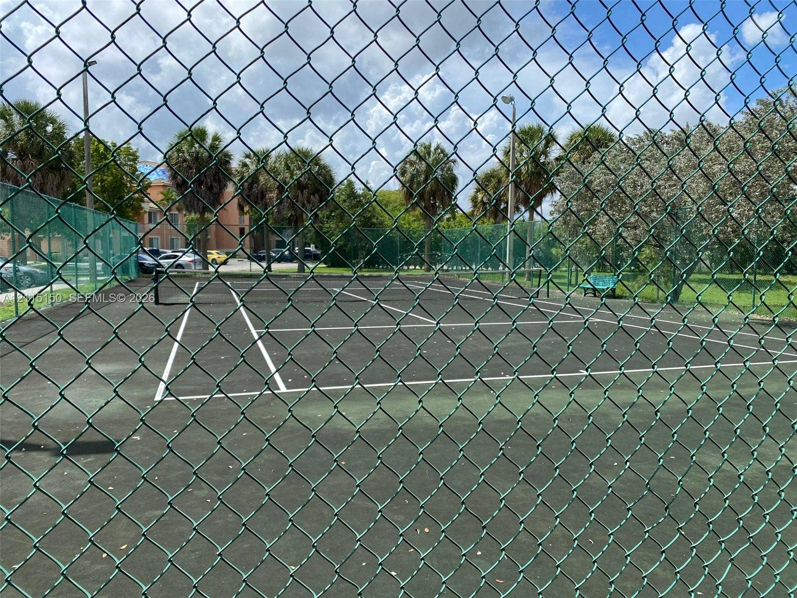 6992 SW 39th Street, #I201, Davie, Davie, FL 33314 Paved tennis court enclosed by chain-link fencing, featuring overhead lighting and perimeter palm trees