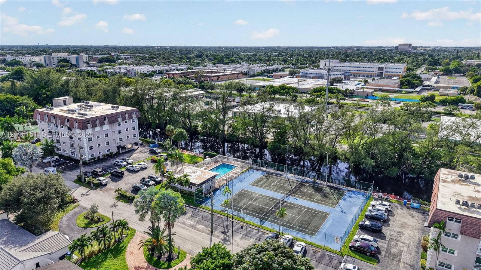 5951 NE 14th Lane, #408N, Fort Lauderdale, FL 33334 Aerial view featuring dual fenced tennis courts, an outdoor swimming pool, mature palm trees, and asphalt parking areas
