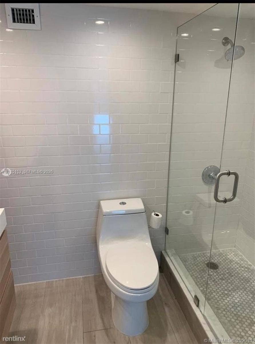 Bathroom featuring a frameless glass shower enclosure, white subway-tiled walls, a dual-flush one-piece toilet, and wood-finish flooring