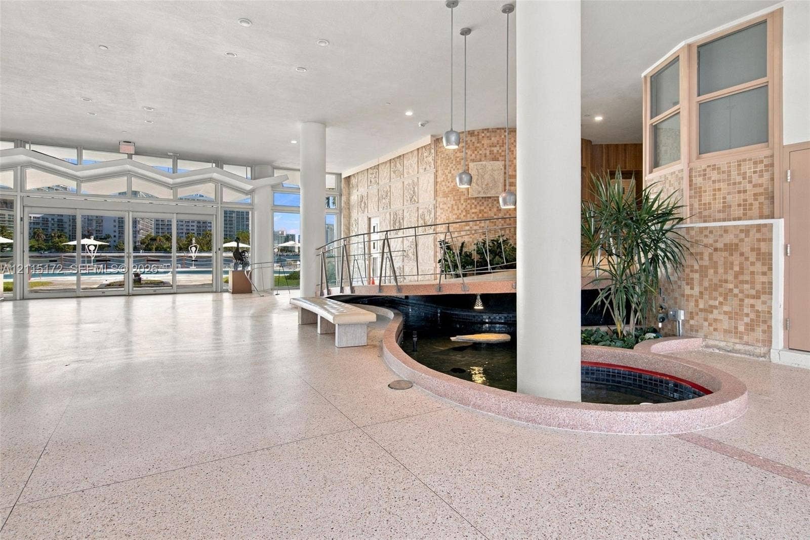 16 Island Avenue, #5C, Miami Beach, FL 33139 Spacious interior featuring terrazzo flooring, a central decorative pond, full-height glass folding doors, structural cylindrical columns, and pendant lighting