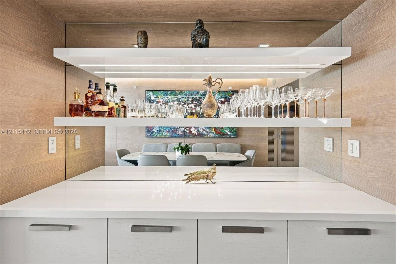 16 Island Avenue, #5C, Miami Beach, FL 33139 Custom built-in bar featuring white cabinetry, polished solid-surface countertop, mirrored backsplash, and tiered floating shelving with integrated LED lighting, framed by wood-finish wall paneling