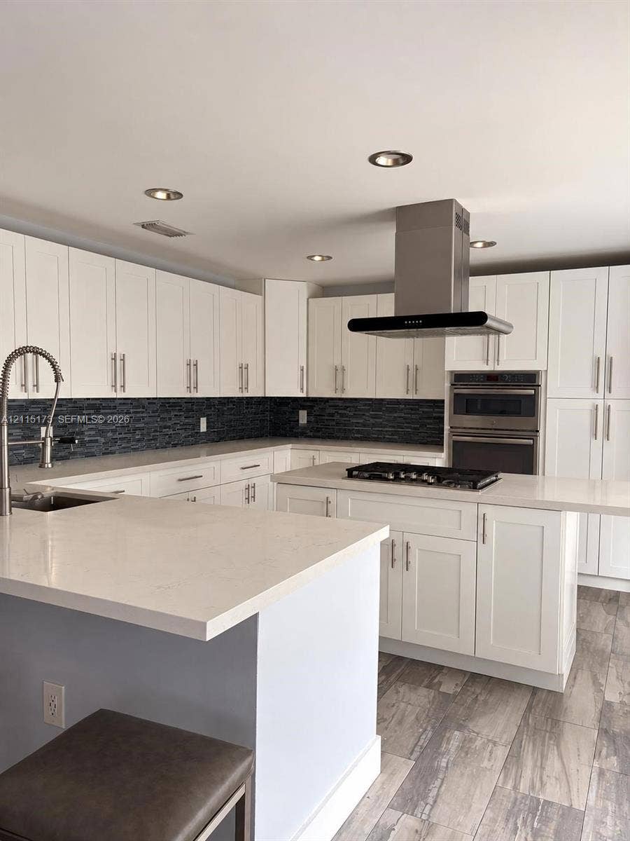 Kitchen featuring white shaker-style cabinetry, quartz countertops, a dark glass tile backsplash, stainless steel built-in double wall ovens with a gas cooktop island, and wood-finish tile flooring