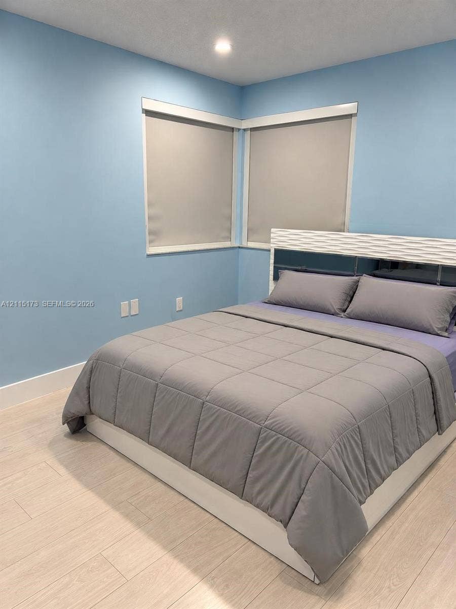 Corner bedroom featuring light-toned wood-finish flooring, recessed ceiling lighting, and dual corner windows with neutral-toned roller shades