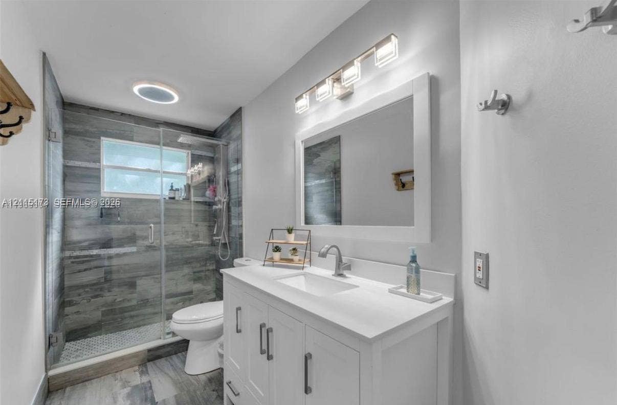 White vanity cabinet with integrated rectangular sink and stone countertop, large framed wall mirror, four-light vanity fixture, and walk-in shower with gray tile surround and glass door