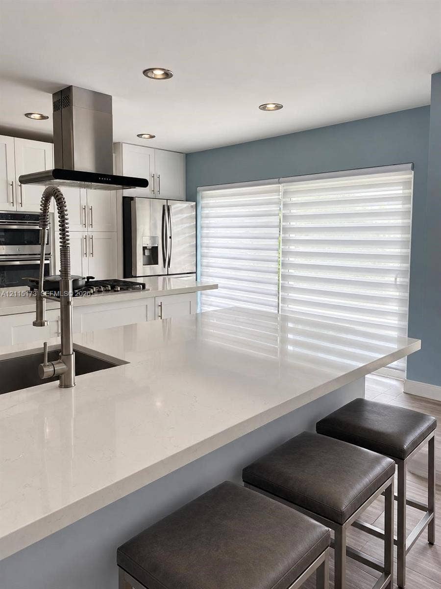 Kitchen featuring a large quartz-topped island with a stainless steel gooseneck faucet, white shaker-style cabinetry, a stainless steel range hood, and recessed ceiling lighting