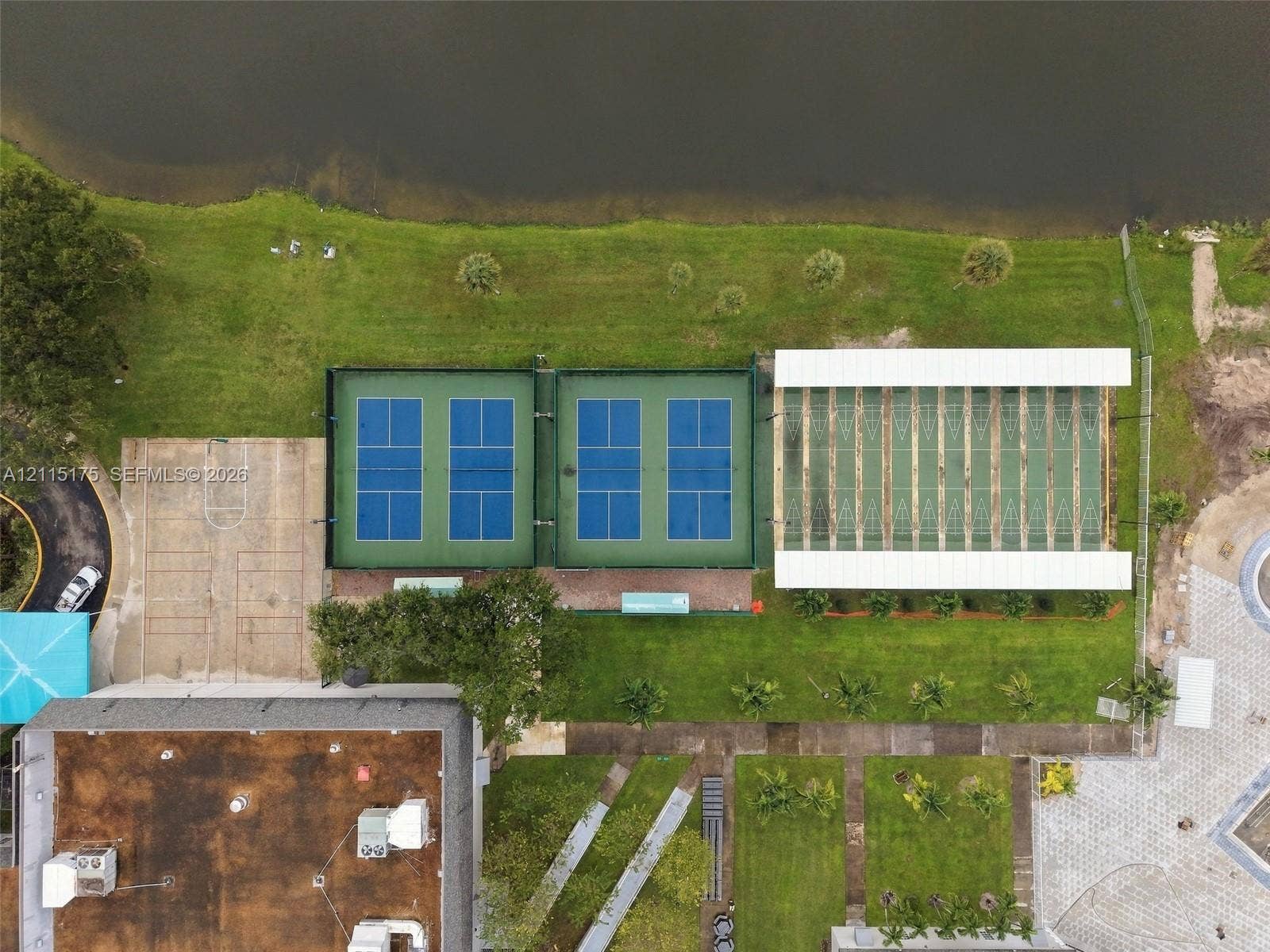 324 Seville N, Delray Beach, FL 33446 Aerial perspective featuring four fenced pickleball courts, a covered multi-lane shuffleboard area, a concrete sport court, and waterfront grounds