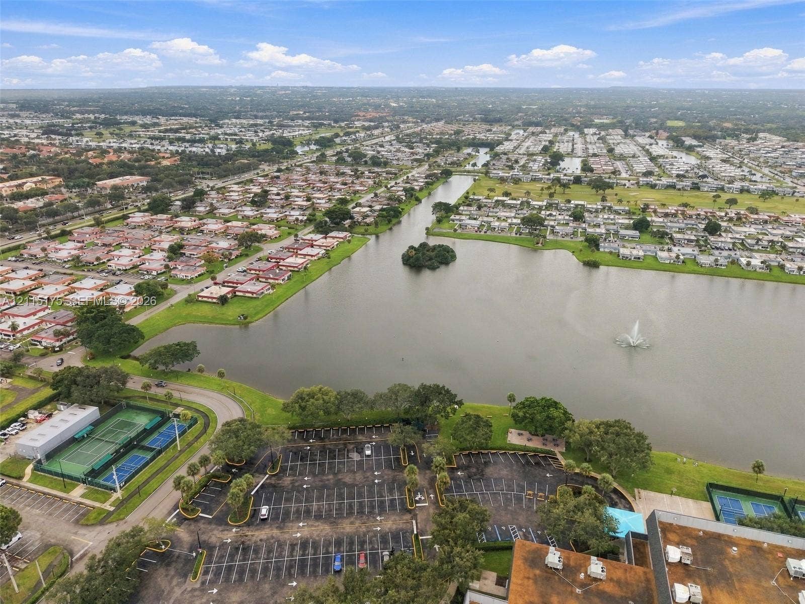 324 Seville N, Delray Beach, FL 33446 Aerial perspective of a lakefront setting featuring a central water fountain, multiple tennis courts, and paved parking areas
