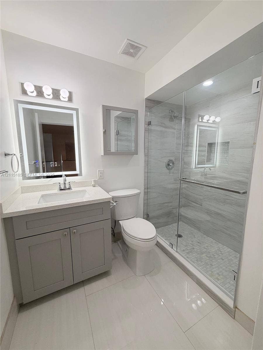 105-107 NE 30th Court, Wilton Manors, FL 33334 Bathroom featuring a vanity with a rectangular sink and stone-finish countertop, backlit mirror, grey cabinetry, and a glass-enclosed shower with tiled walls and flooring