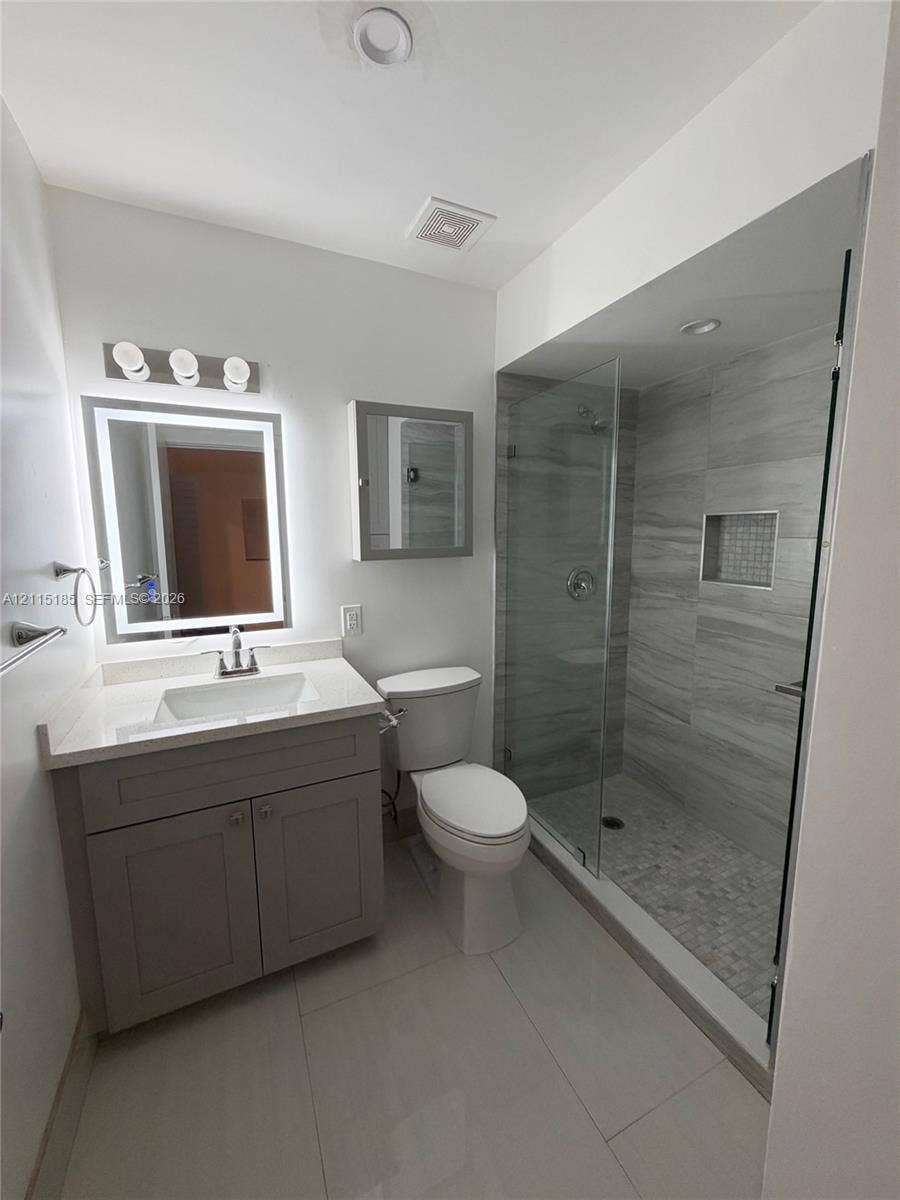 105-107 NE 30th Court, Wilton Manors, FL 33334 Bathroom featuring a vanity with white countertop, integrated sink, and illuminated mirror, paired with a walk-in shower encased in glass with stone-finish wall tiling and a built-in recessed niche