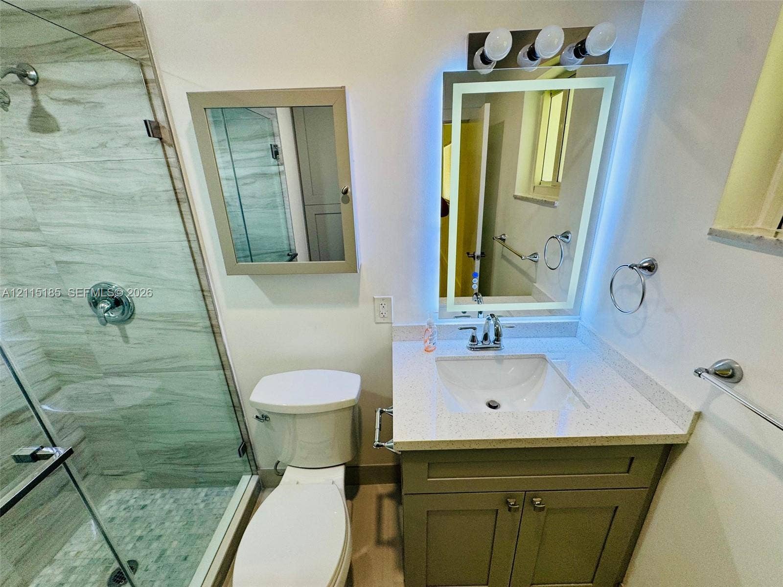 105-107 NE 30th Court, Wilton Manors, FL 33334 Bathroom featuring a vanity with a light-toned countertop, integrated basin, illuminated mirror with integrated lighting, a walk-in shower with stone-finish tile surround and pebble tile flooring, and a glass shower enclosure