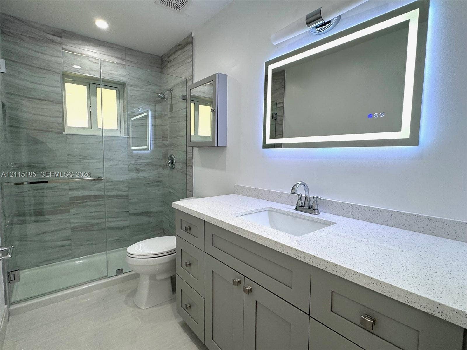 105-107 NE 30th Court, Wilton Manors, FL 33334 Bathroom featuring a gray shaker-style vanity with a light-toned quartz countertop, integrated LED-backlit mirror, and walk-in shower enclosed by glass paneling with full-height stone-finish tile surround