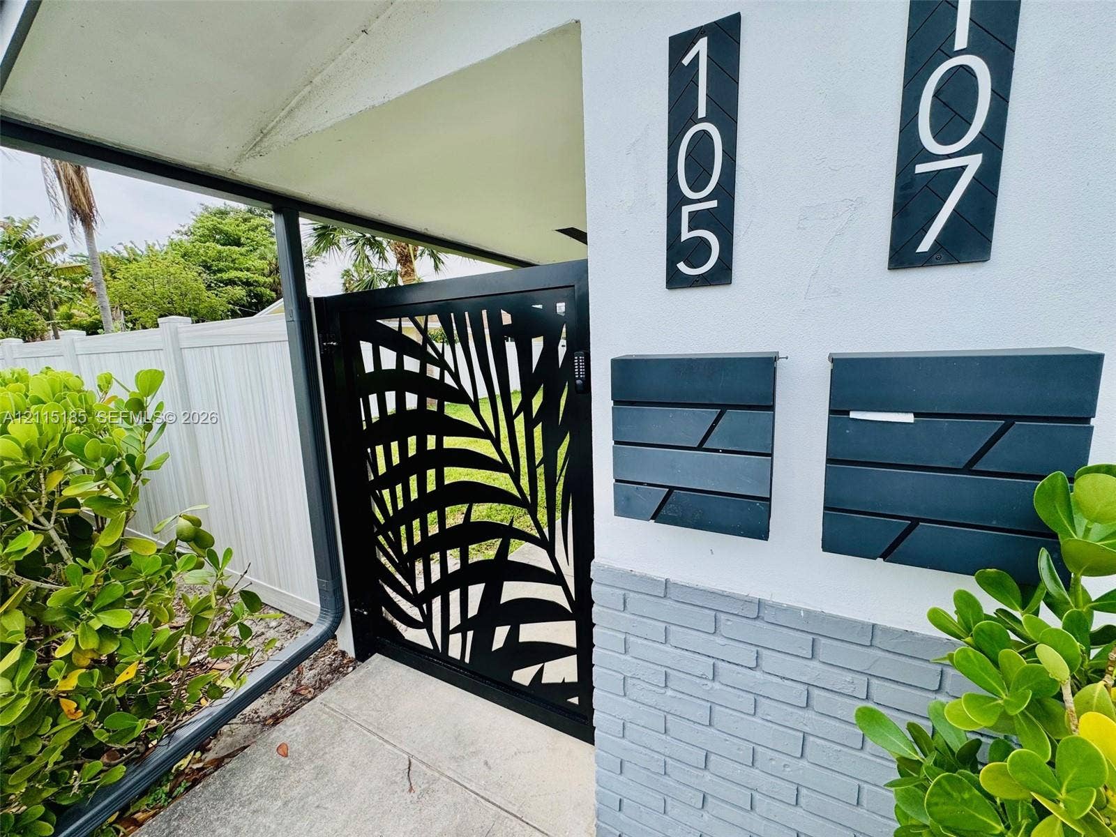 105-107 NE 30th Court, Wilton Manors, FL 33334 Black metal decorative gate with palm leaf pattern, wall-mounted matte black mailboxes, vertical modern address signage, and grey brick exterior accent