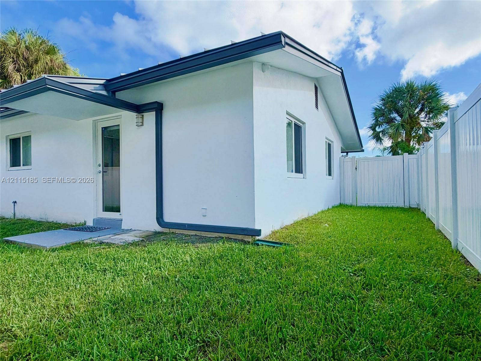 105-107 NE 30th Court, Wilton Manors, FL 33334 Exterior facade featuring white stucco finish, dark-trimmed eaves, a glass-paneled exterior door, and a white vinyl privacy fence enclosing the lawn
