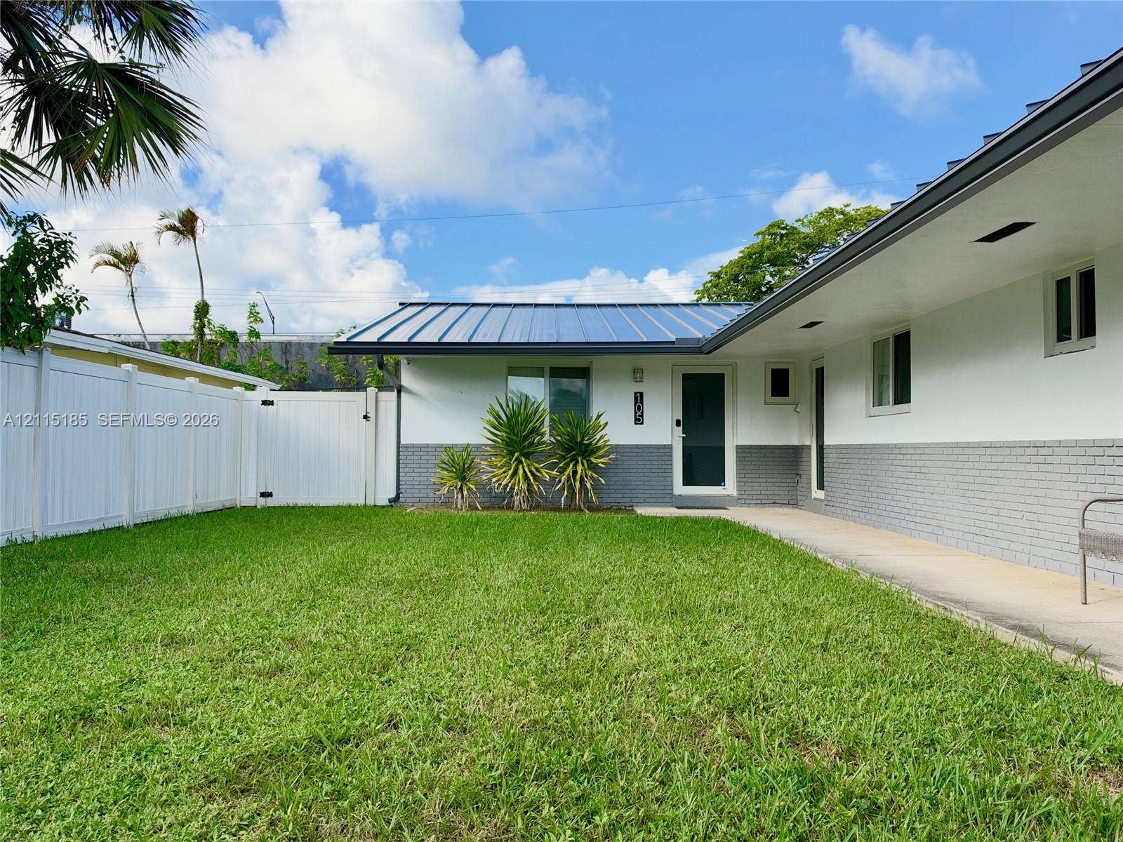 105-107 NE 30th Court, Wilton Manors, FL 33334 Exterior facade featuring a standing-seam metal roof, white stucco walls with grey brick lower siding, and a white vinyl privacy fence enclosing a grass lawn