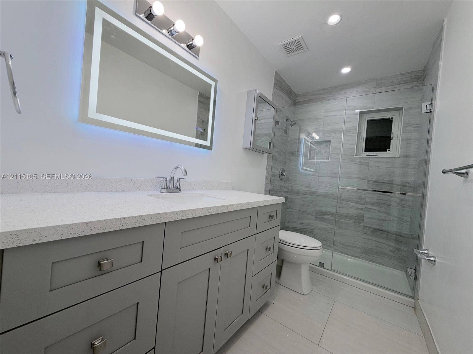 105-107 NE 30th Court, Wilton Manors, FL 33334 Grey shaker-style vanity with a light-toned solid surface countertop, rectangular undermount sink, illuminated mirror, and glass-enclosed shower featuring horizontal-pattern wall tiling