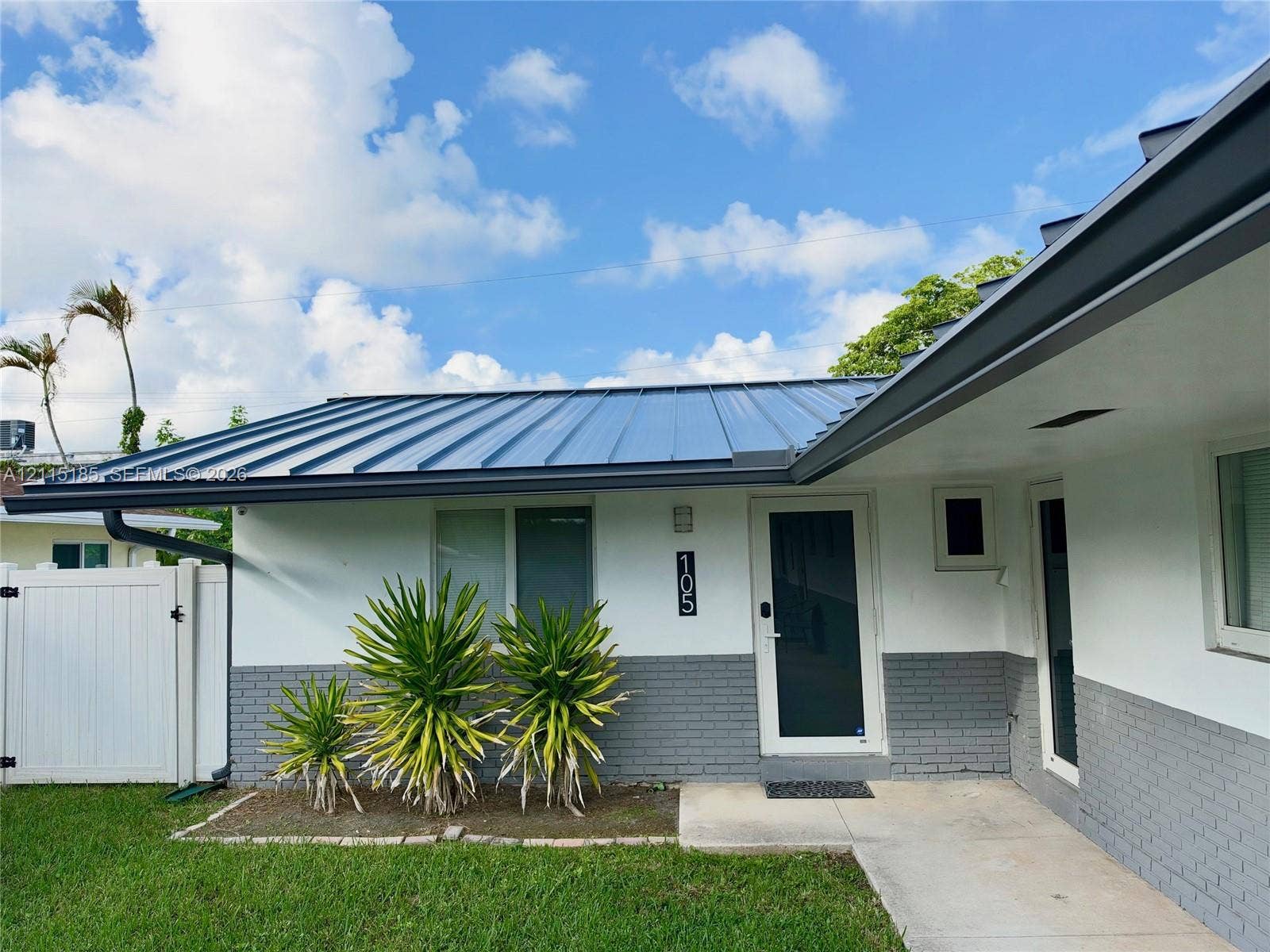 105-107 NE 30th Court, Wilton Manors, FL 33334 Exterior facade featuring a metal roof, two-tone painted brick and smooth finish siding, concrete entryway, and white vinyl privacy fencing