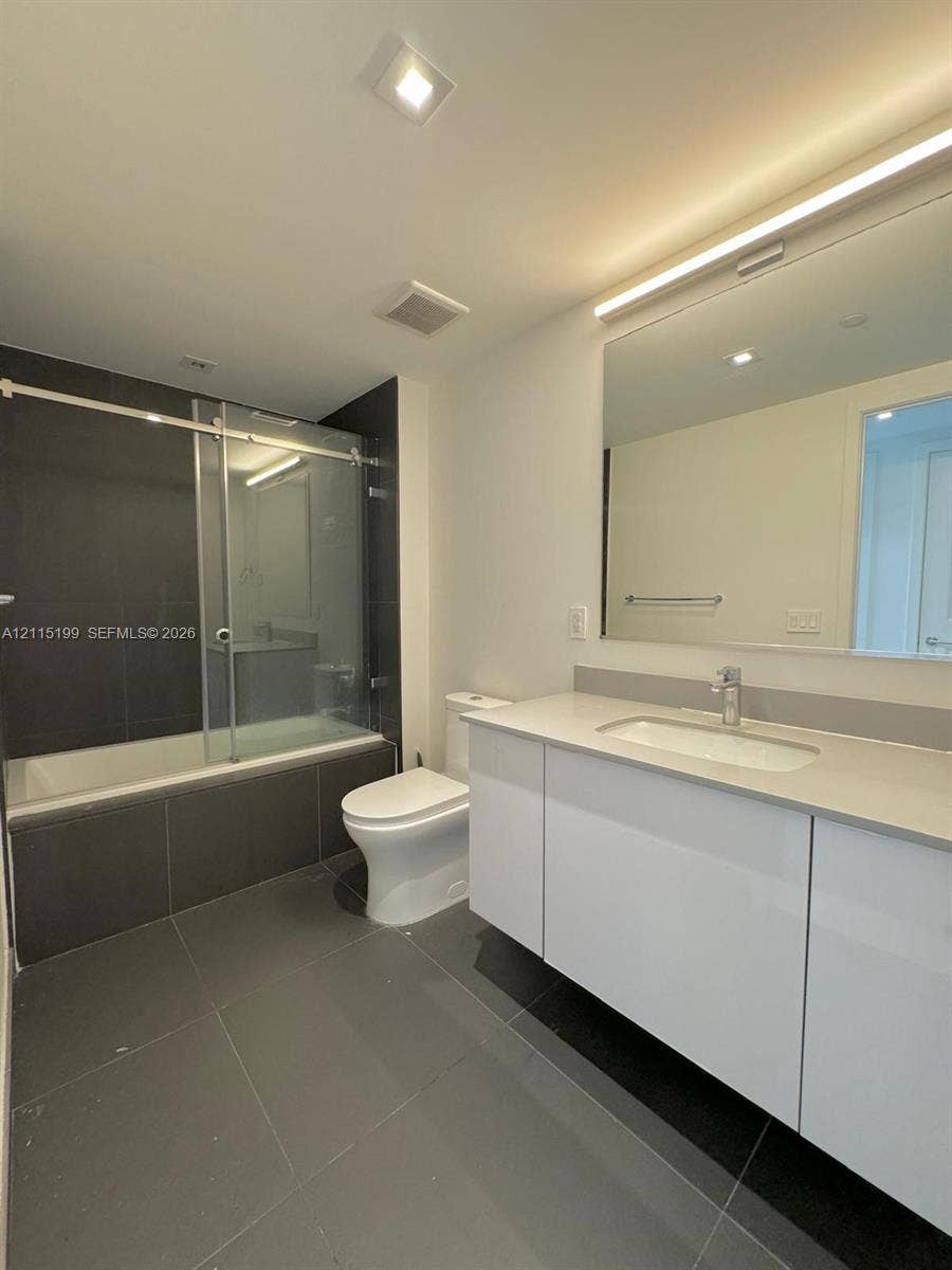 1600 NE 1st Avenue, #3120, Miami, FL 33132 Bathroom featuring a white vanity with a neutral solid-surface countertop, an integrated undermount sink, a large wall-mounted mirror with integrated lighting, a soaking tub with a glass sliding enclosure, and large-format gray tile flooring