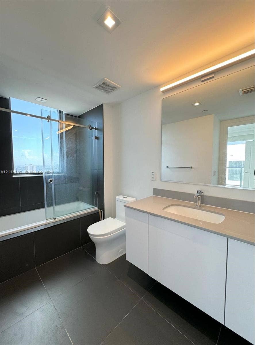 1600 NE 1st Avenue, #3120, Miami, FL 33132 Bathroom featuring a white flat-panel vanity with an integrated countertop, large wall-mounted mirror with integrated lighting, oversized charcoal floor tile, and a soaking tub with a sliding glass enclosure and charcoal tile surround