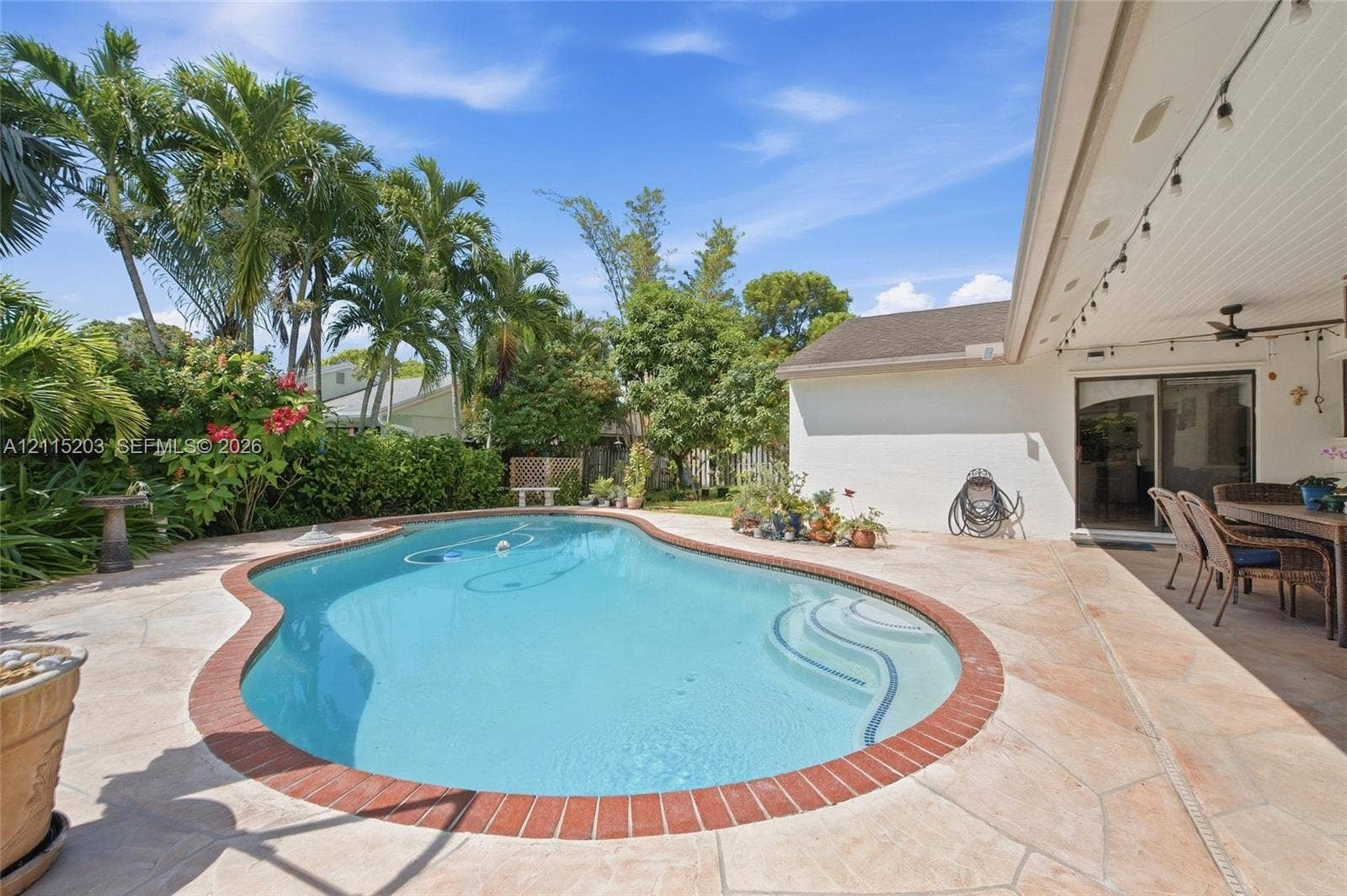 Free-form swimming pool with integrated steps, surrounded by natural stone pavers and a brick coping border, featuring a covered patio and tropical landscaping