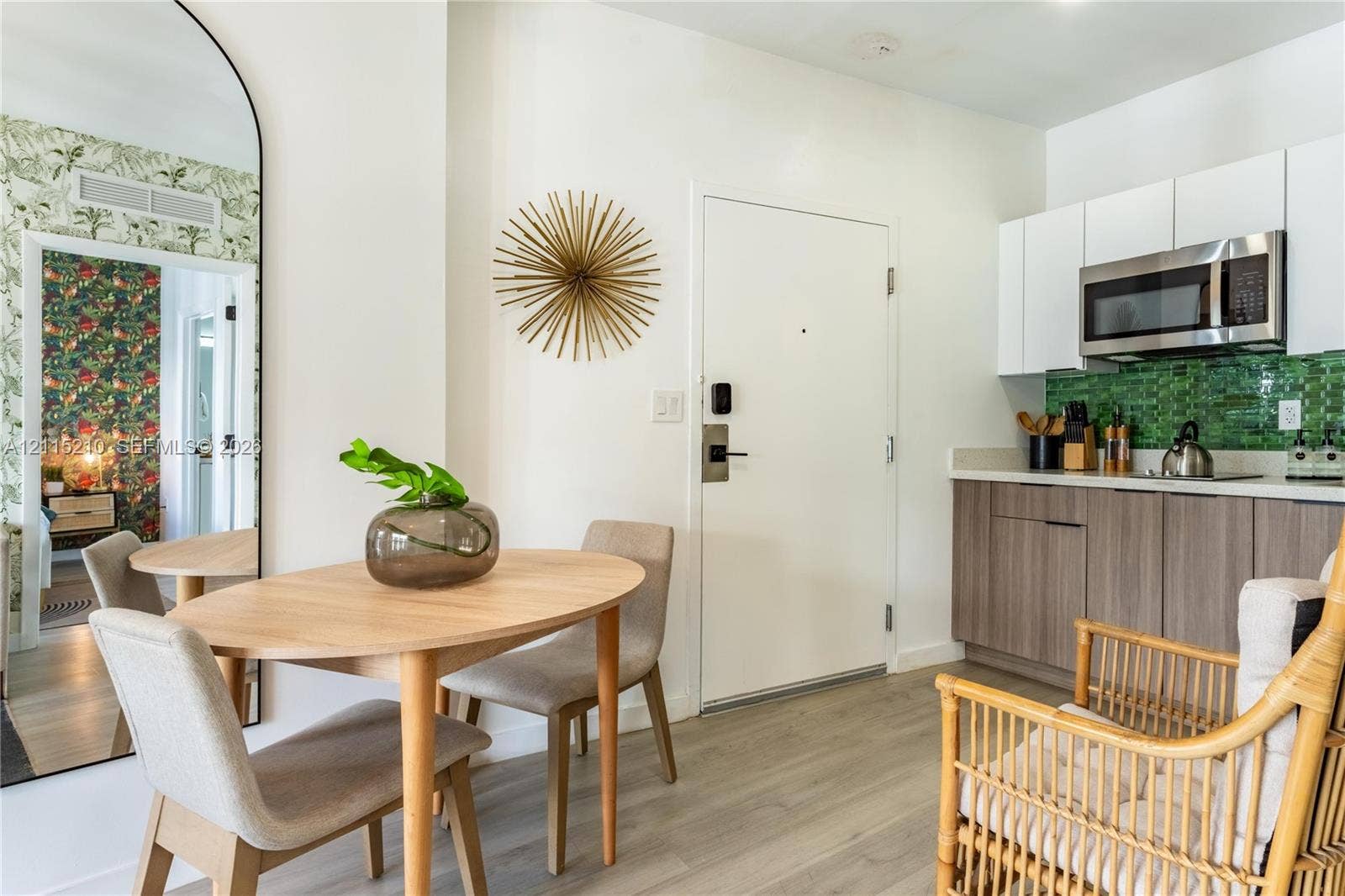136 NW 26th Street, #507, Miami, FL 33127 Kitchen featuring wood-finish lower cabinetry, white upper cabinetry, integrated stainless steel microwave, and green glass tile backsplash, accented by wood-finish flooring and a full-length arched wall mirror