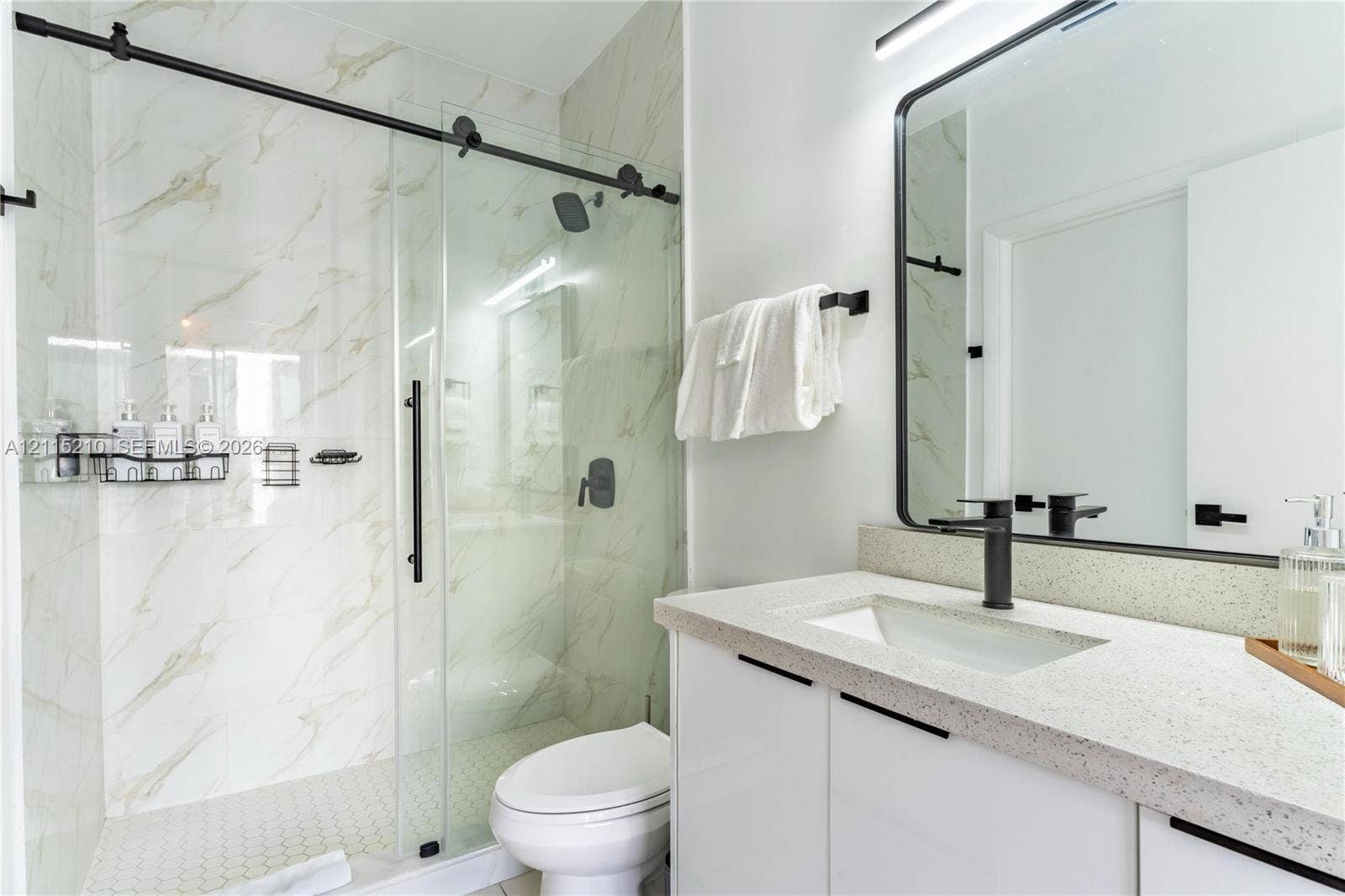 136 NW 26th Street, #507, Miami, FL 33127 Walk-in shower with sliding glass door and stone-patterned wall tile, vanity with speckled countertop, rectangular undermount sink, matte black fixtures, and framed vanity mirror