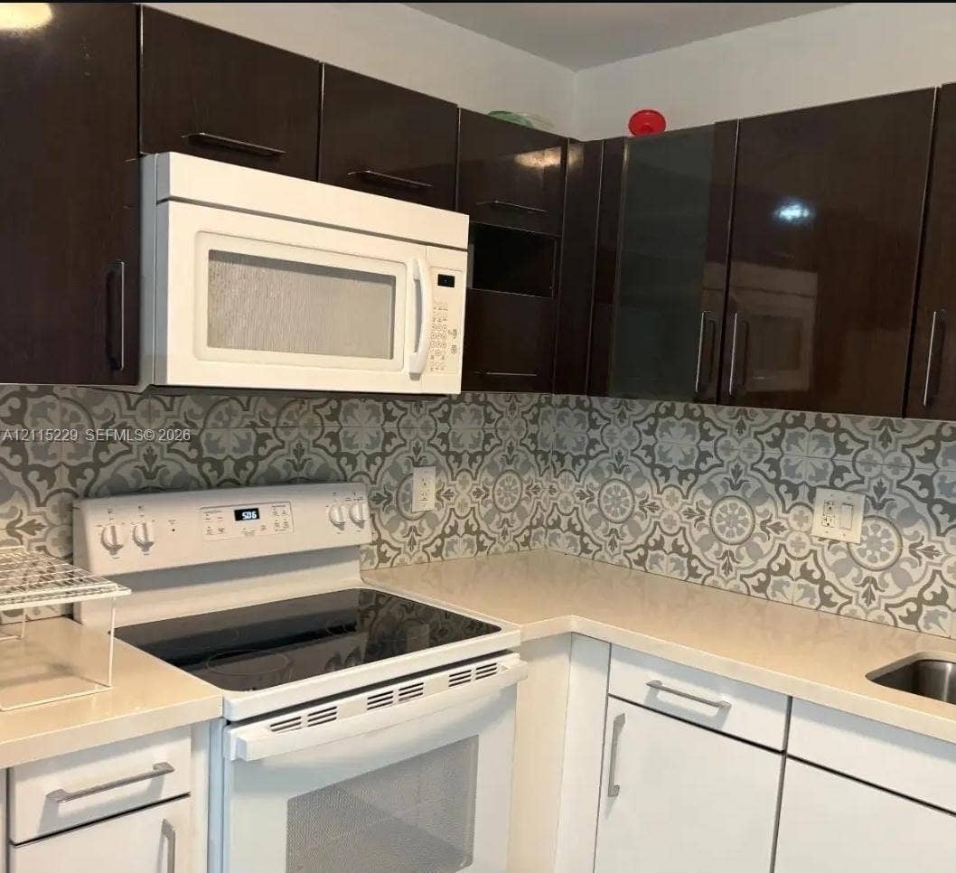 2020 NE 135th Street, #211, North Miami, FL 33181 Kitchen featuring dark-toned upper cabinetry, white lower cabinetry with modern hardware, patterned tile backsplash, solid-surface countertops, and a white over-the-range microwave