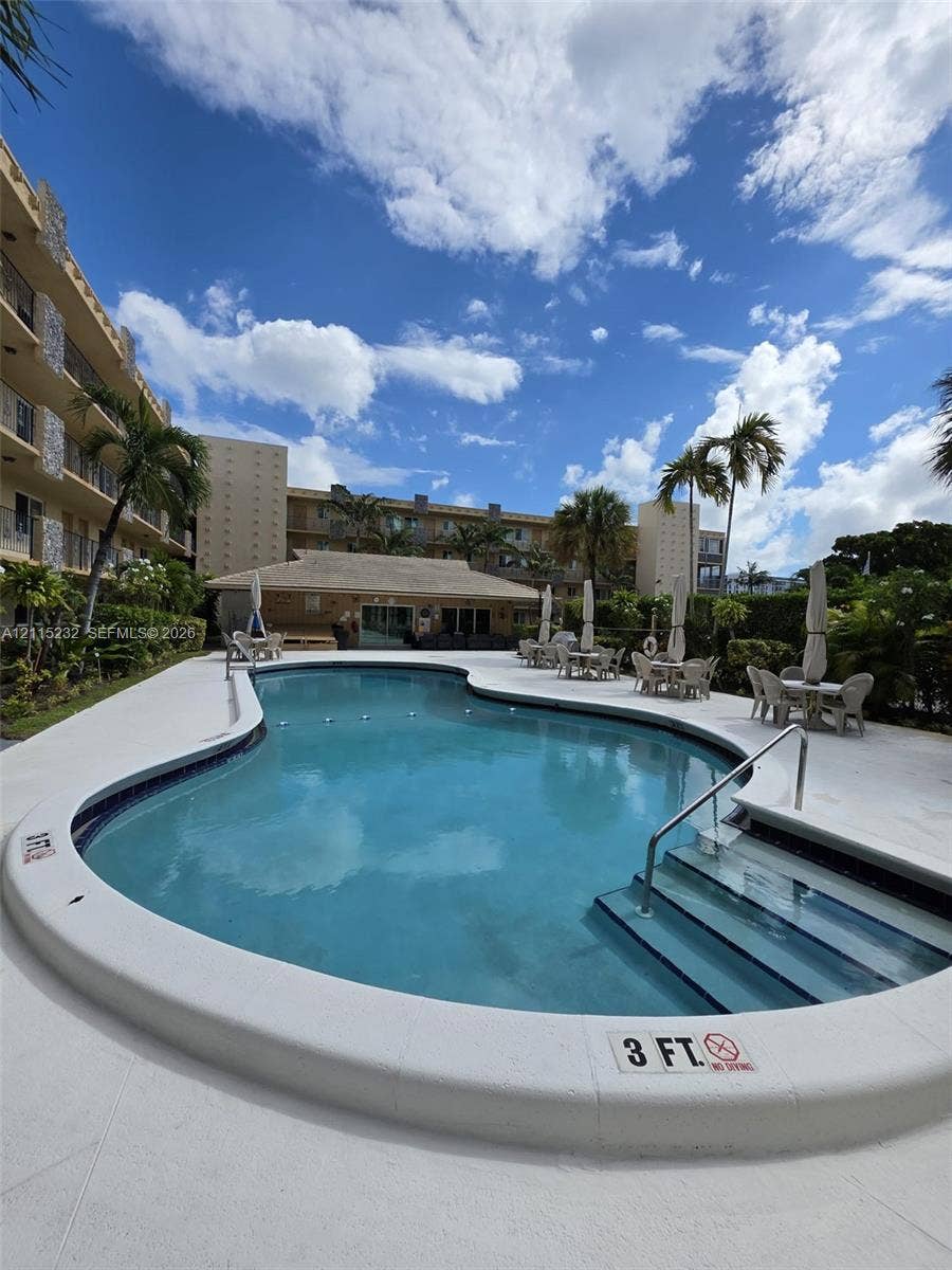Curved swimming pool featuring a concrete surround, built-in steps with metal handrails, a poolside structure, and mature palm trees