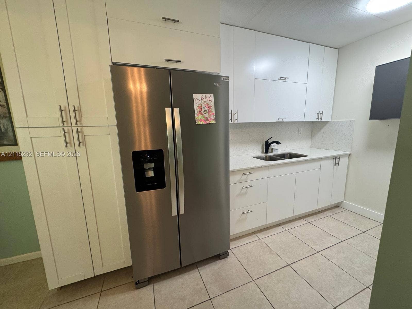 Kitchen featuring white shaker-style cabinetry, a stainless steel side-by-side refrigerator, solid-surface countertops, dual-basin stainless steel sink with matte black faucet, and tiled flooring