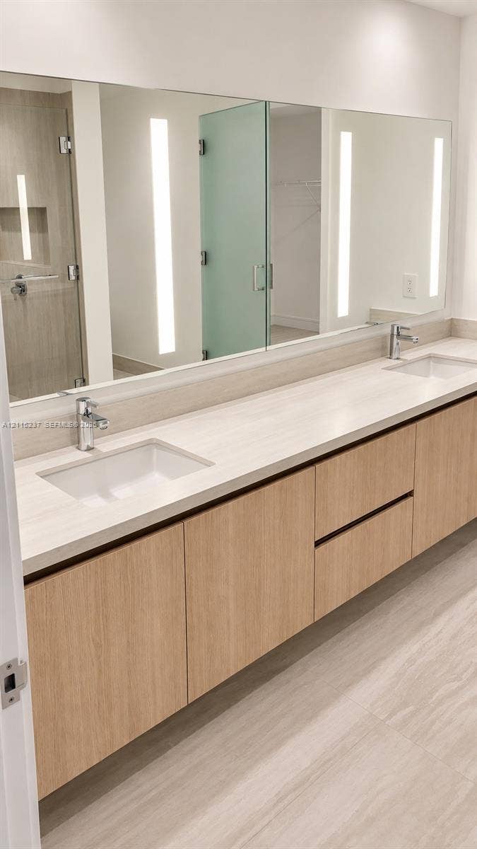 2411 Laguna Circle, #802, North Miami, North Miami, FL 33181 Floating wood-finish vanity with dual integrated rectangular sinks, light-toned stone-finish countertop, integrated linear vertical LED mirror lighting, and polished chrome fixtures