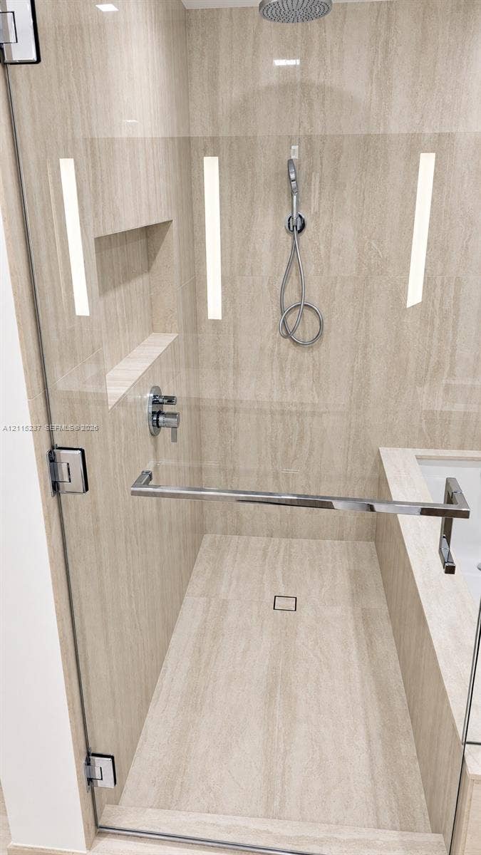 2411 Laguna Circle, #802, North Miami, North Miami, FL 33181 Walk-in shower featuring natural-stone tile surrounds, a rain-style showerhead with a handheld attachment, a built-in recessed niche, and a frameless glass enclosure with a metallic towel bar